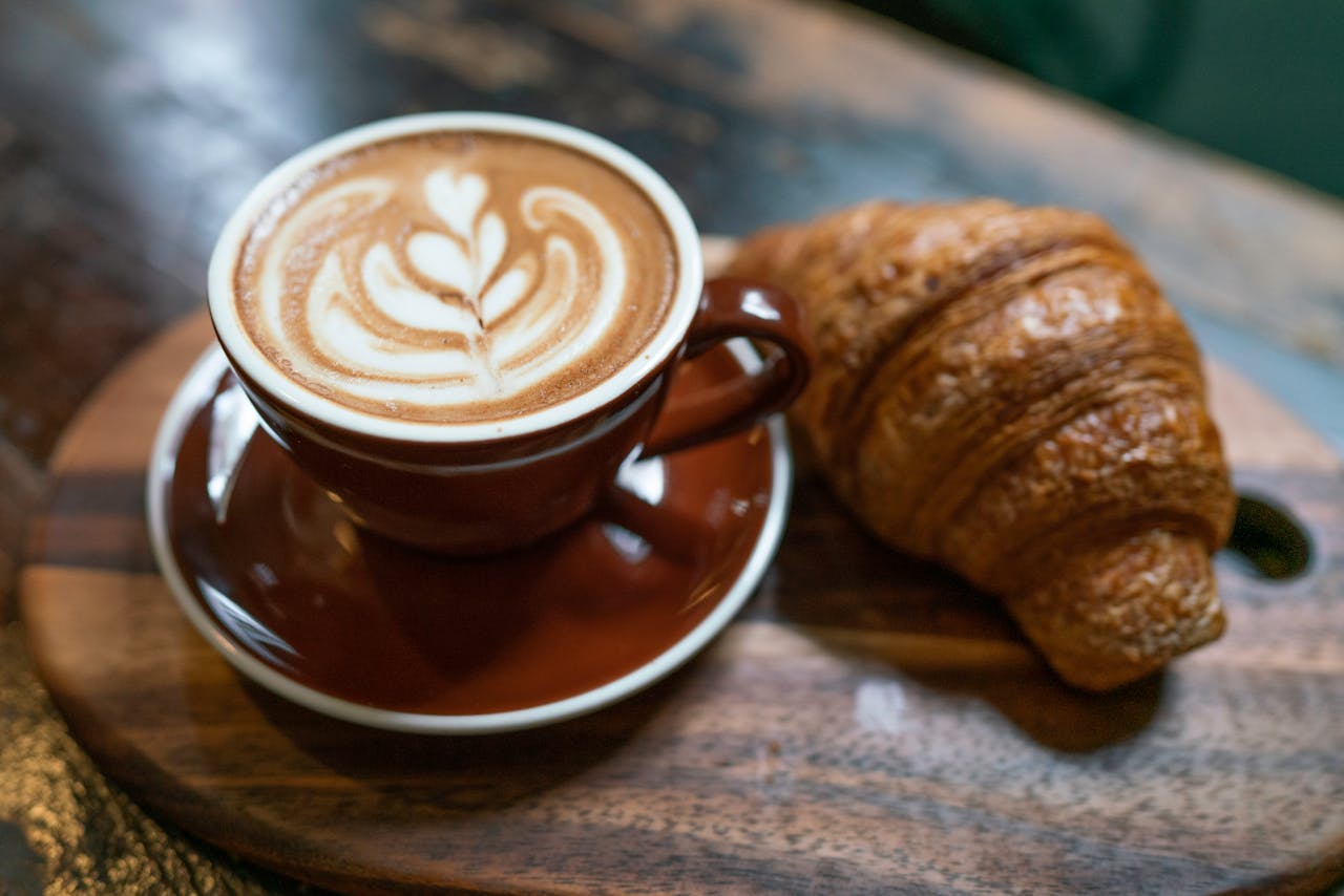 A delicious cappuccino with latte art served with a fresh croissant on a wooden tray.